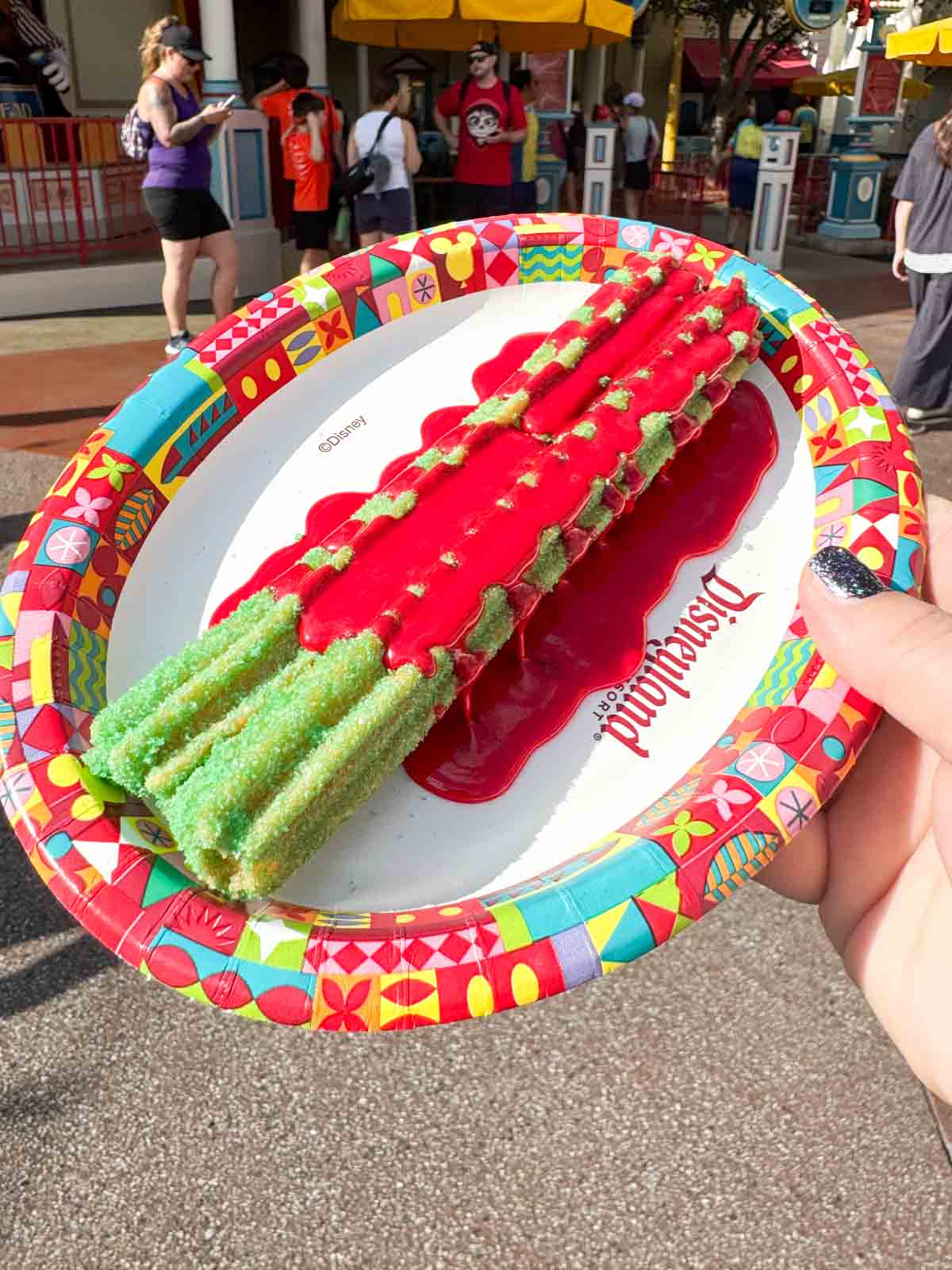 hand holding a plate with a green poison apple churro on top